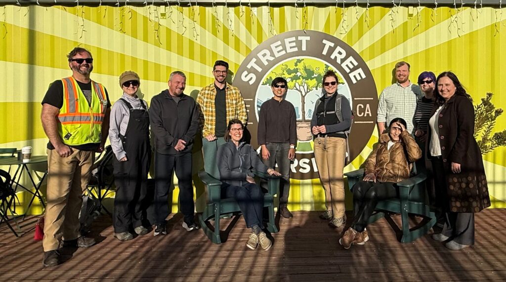 A group of eleven people posing for a photo in front of a bright yellow mural with a circular Street Tree sign in the center.